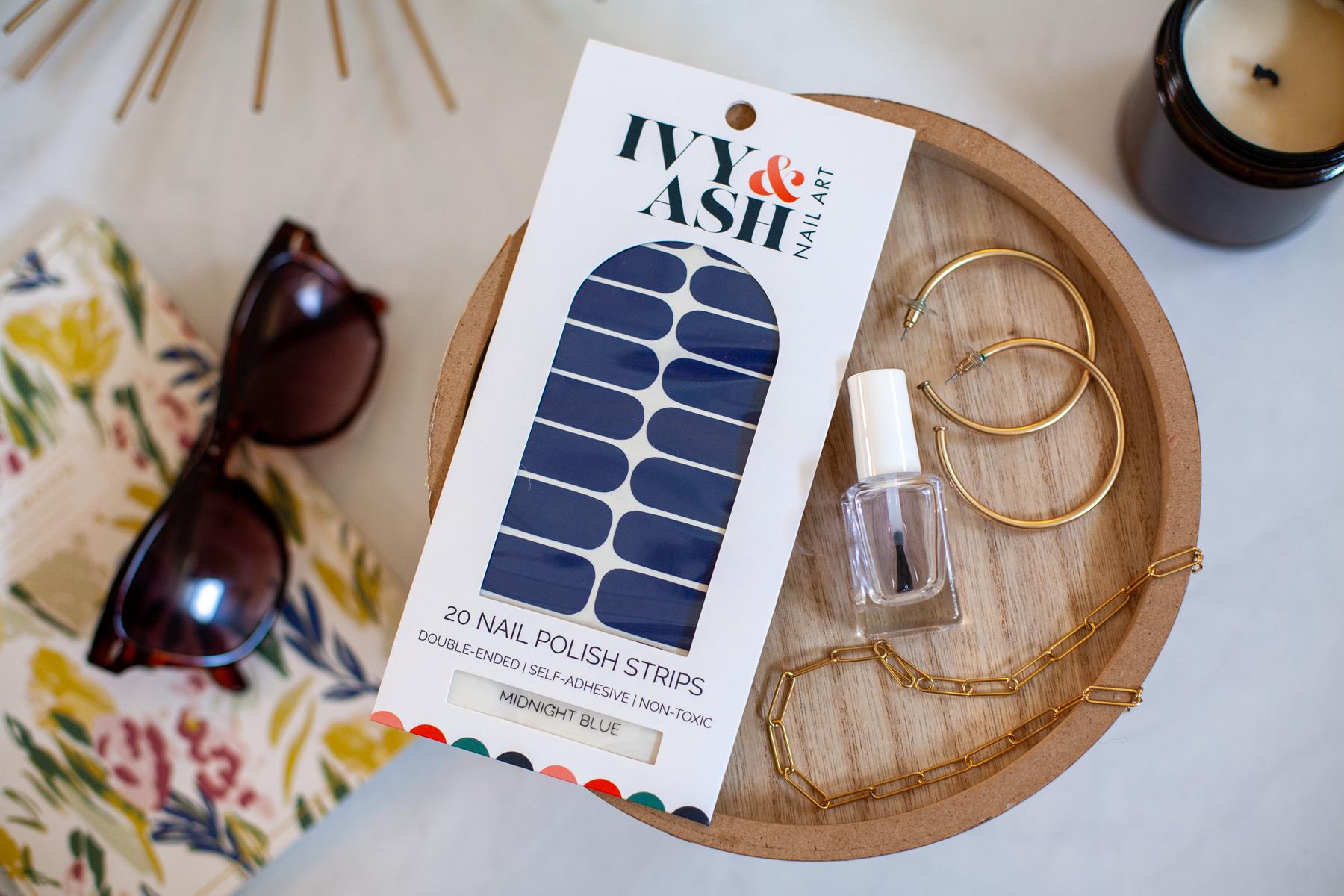 A package of Ivy & Ash Nail Art double-ended self-adhesive nail polish strips in Midnight Blue, styled on a round wooden tray with gold hoop earrings, a small bottle of nail polish, sunglasses, and a candle.